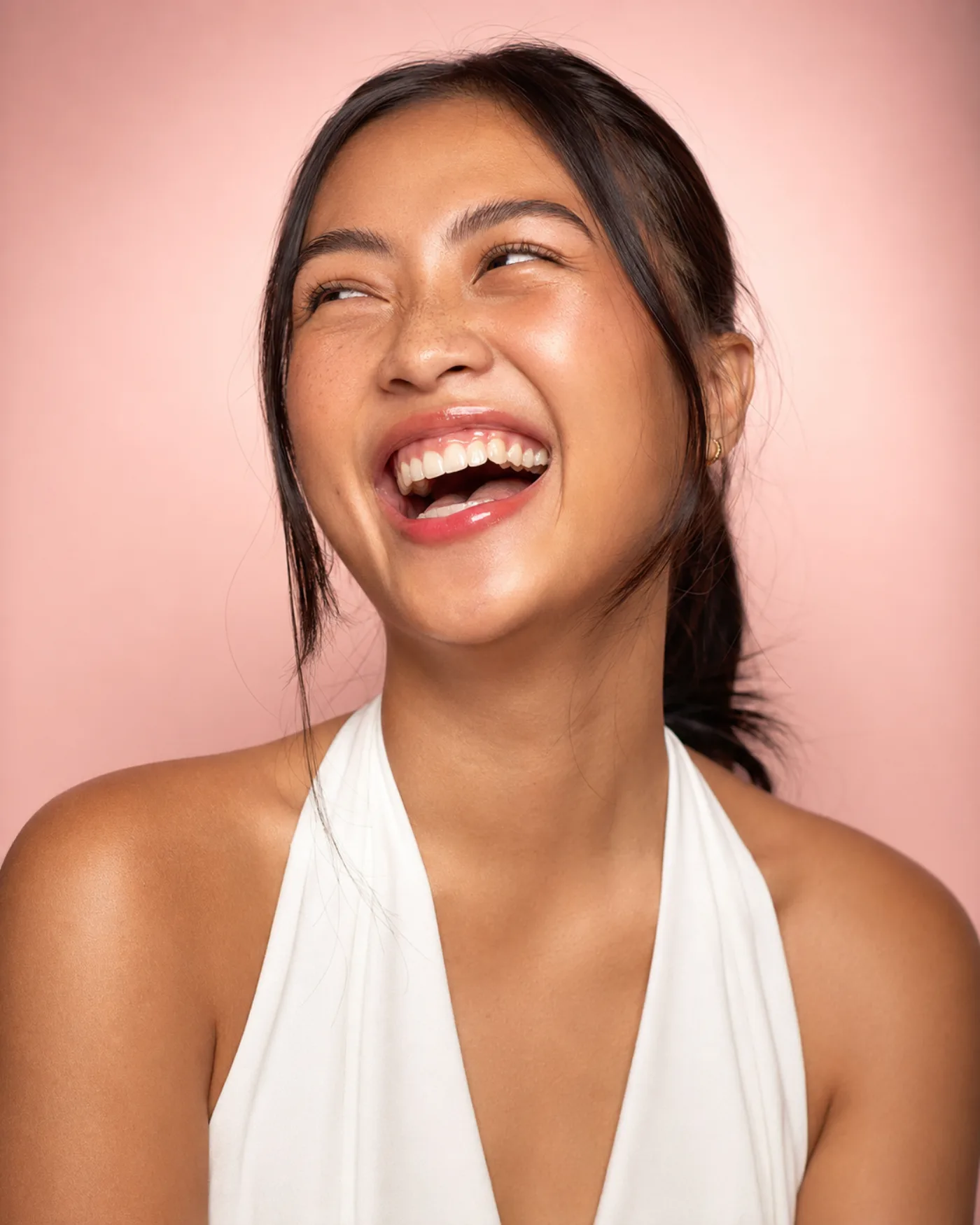 Waist-up studio portrait of a laughing dark-haired woman in a white halter top against a pink wall, eyes creased shut and freckles scattered across her nose