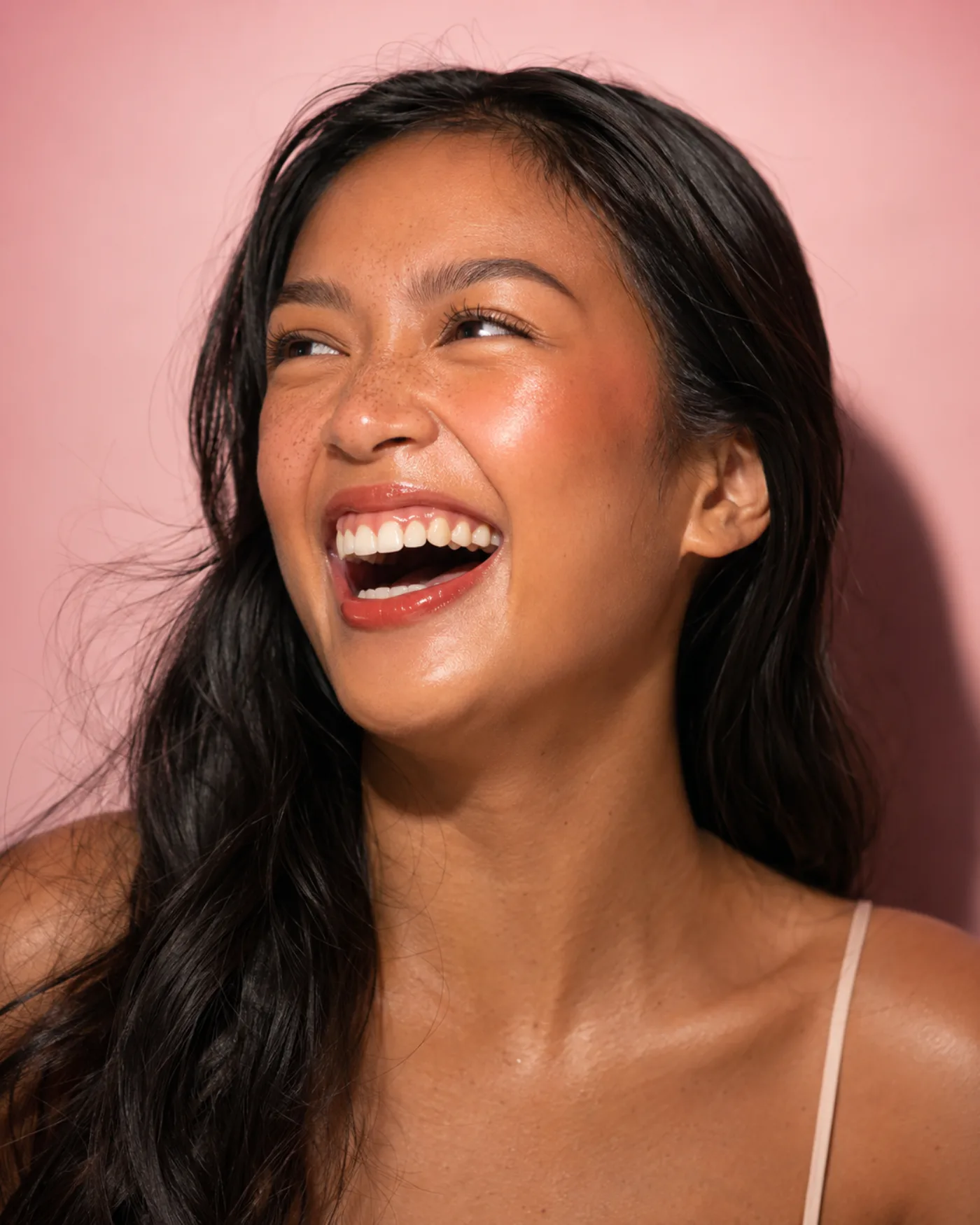 Studio portrait of a laughing woman with long dark hair falling over one shoulder against a pink wall, warm blush high on the cheek and a thin strap at her shoulder